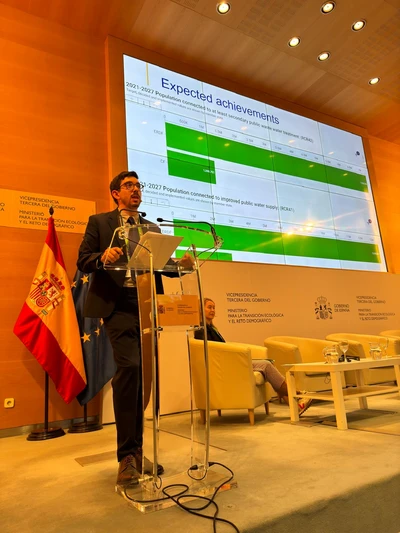 Ramses steht am Rednerpult vor einer Präsentationsfolie mit grün-weißer Grafik und Text 'Expected achievements'. Im Hintergrund spanische und EU-Flagge, gelbe Sessel und Tisch mit Gläsern.