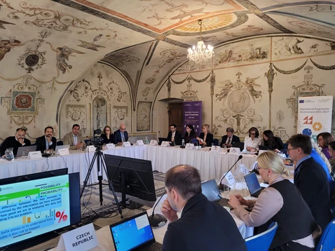 Konferenzraum mit barockem Deckengemälde und Kronleuchter, Teilnehmer sitzen an U-förmigem Tisch mit Namensschildern, zwei Bildschirme und Kamera in der Mitte.