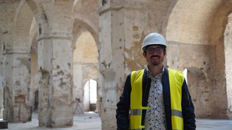 Ramses steht mit Bauhelm und gelber Warnweste in einem historischen, steinernen Gewölbe mit hohen Rundbögen.